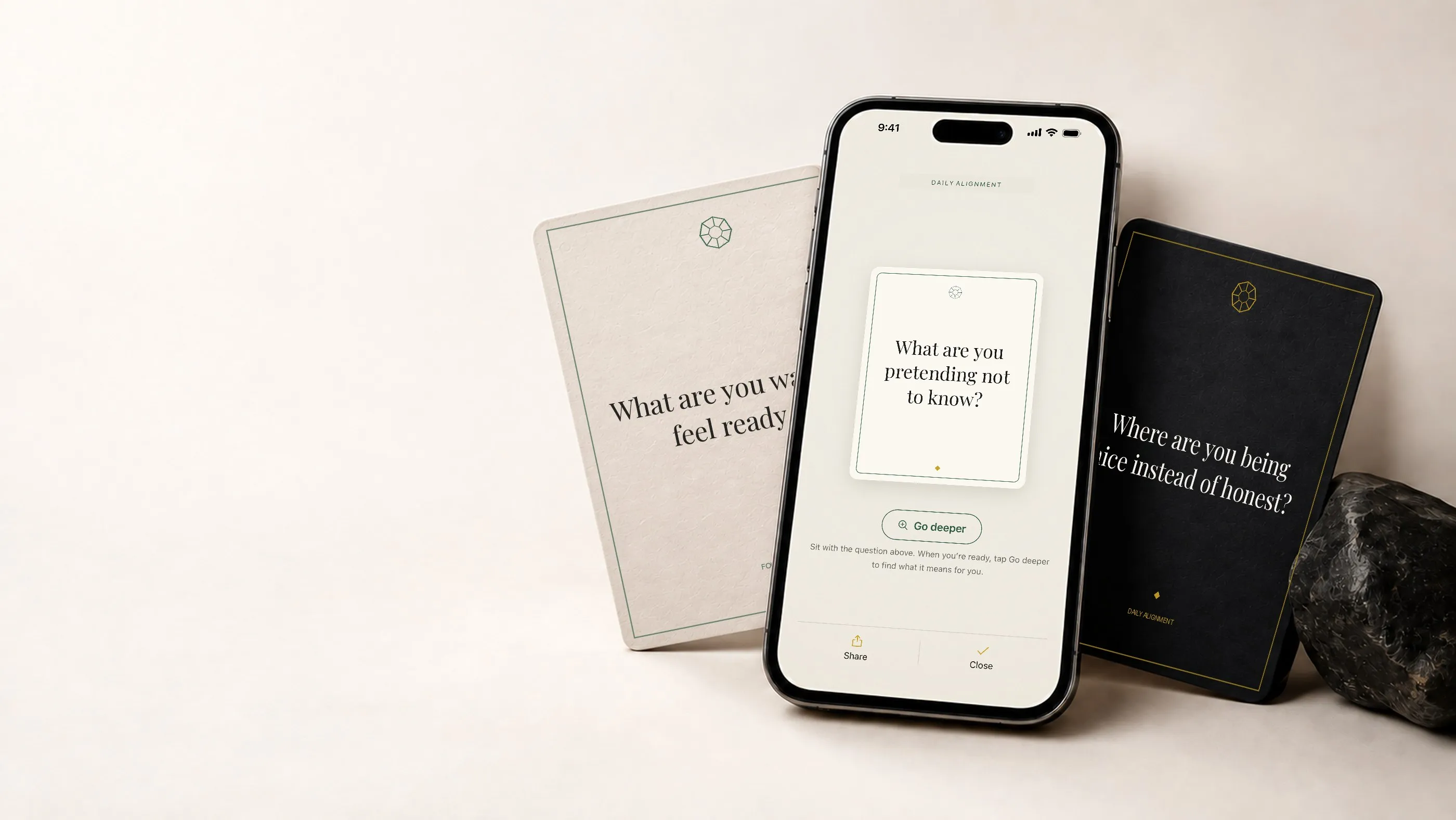 A phone beside a dark stone showing the question, "What are you pretending not to know?", between two cards reading "What are you waiting to feel ready for?" and "Where are you being nice instead of honest?"